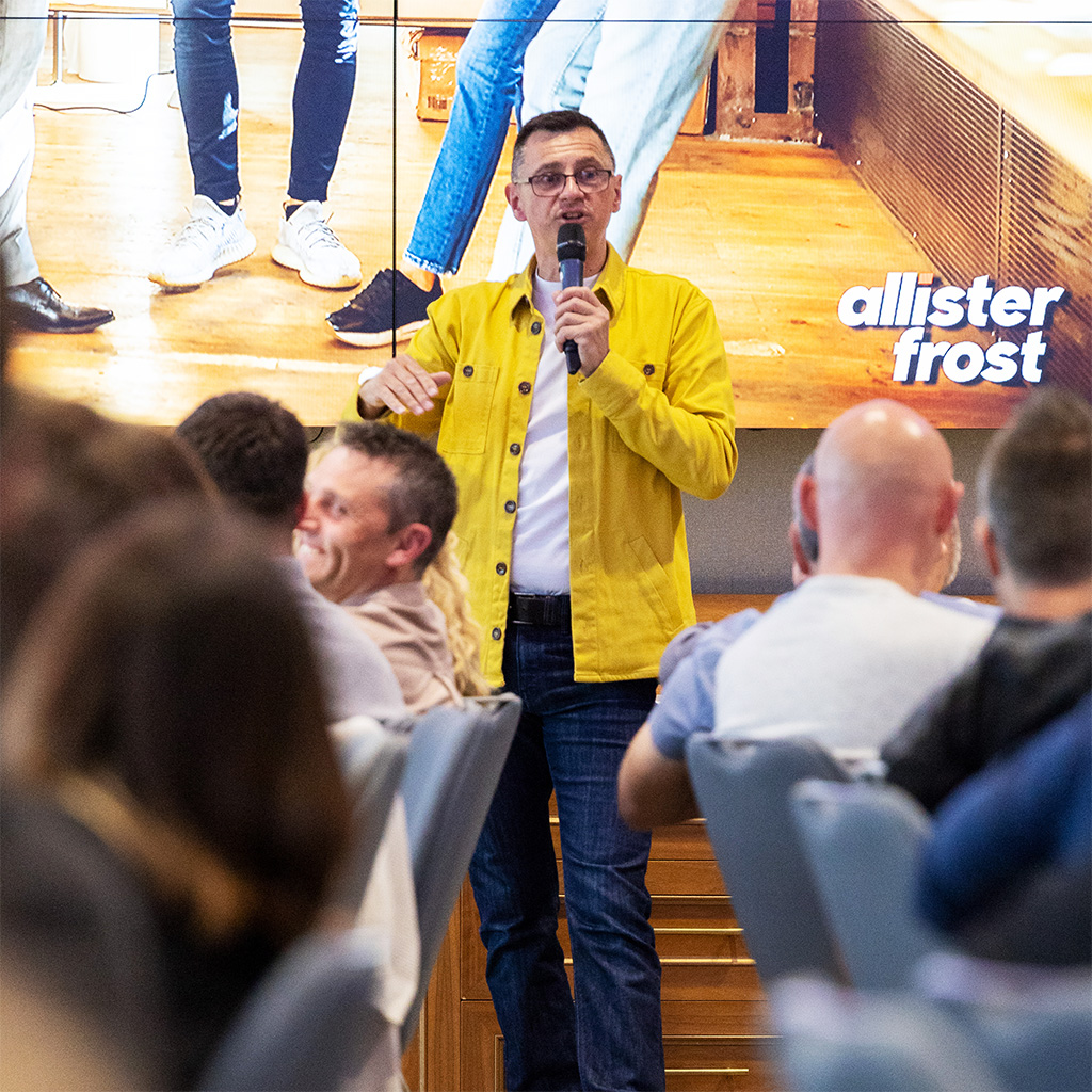 Allister Frost in yellow overshirt speaking into a microphone in front of an audience